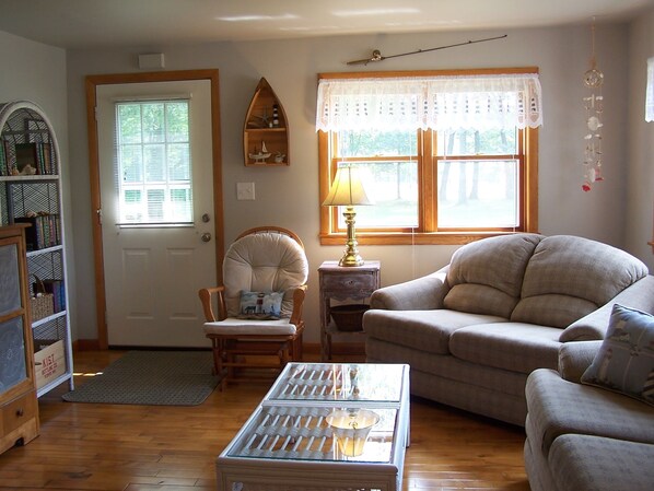 Un salón acogedor con suelo de madera, un sofá color beige, un sillón blanco, una mesita y una estantería.