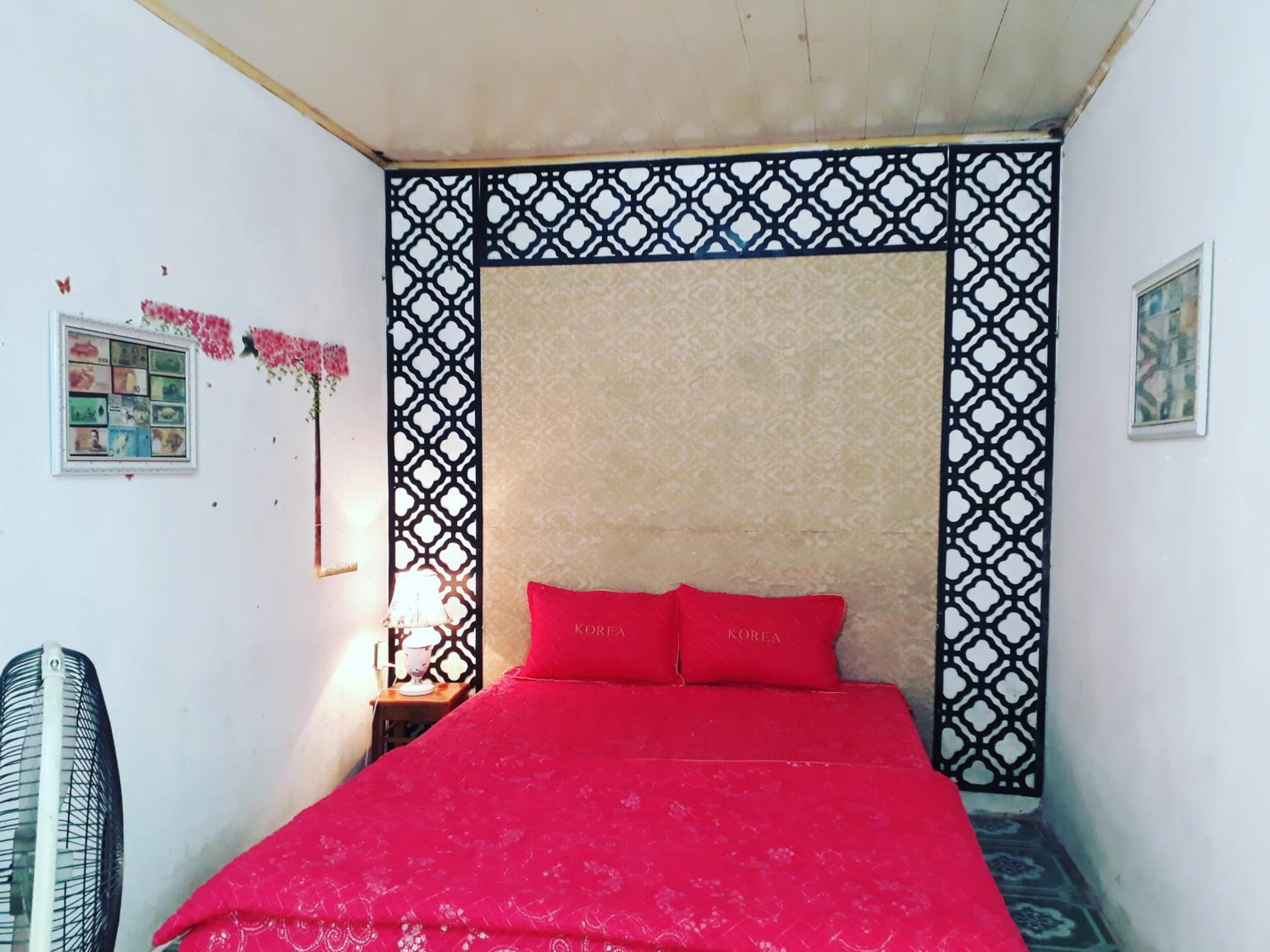 Ein Zimmer mit einem Bett, einem Wandventilator, einem dekorativen Raumteiler und einem kleinen Tisch mit einer Lampe.