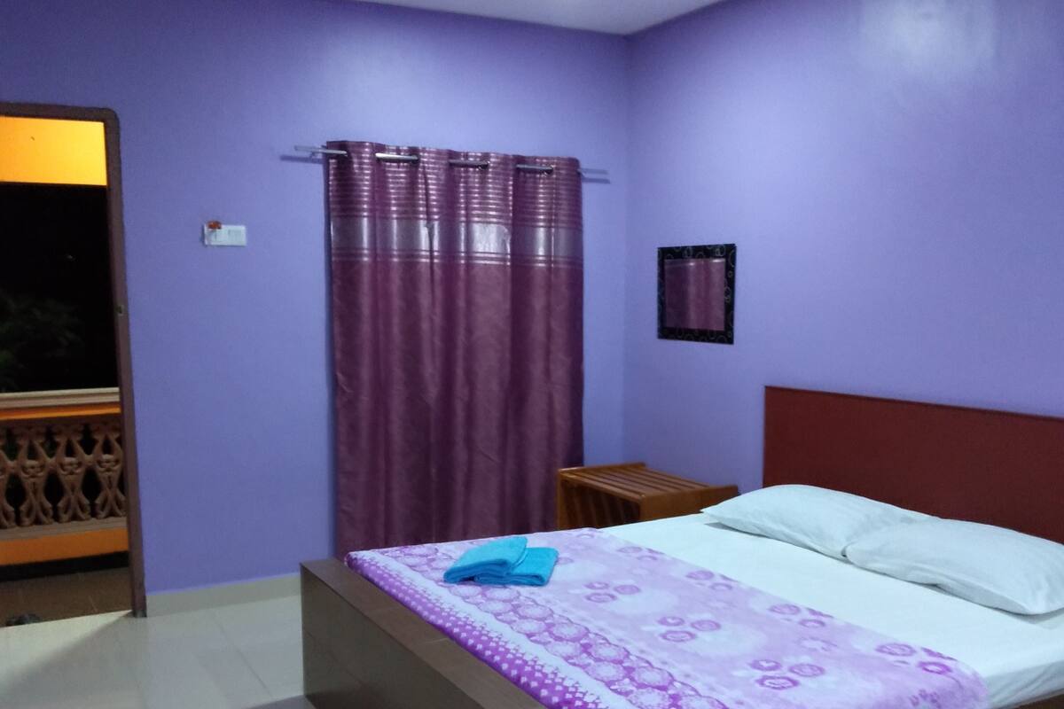 A bedroom with a bed, a nightstand, a window with curtains, and a wall-mounted light.