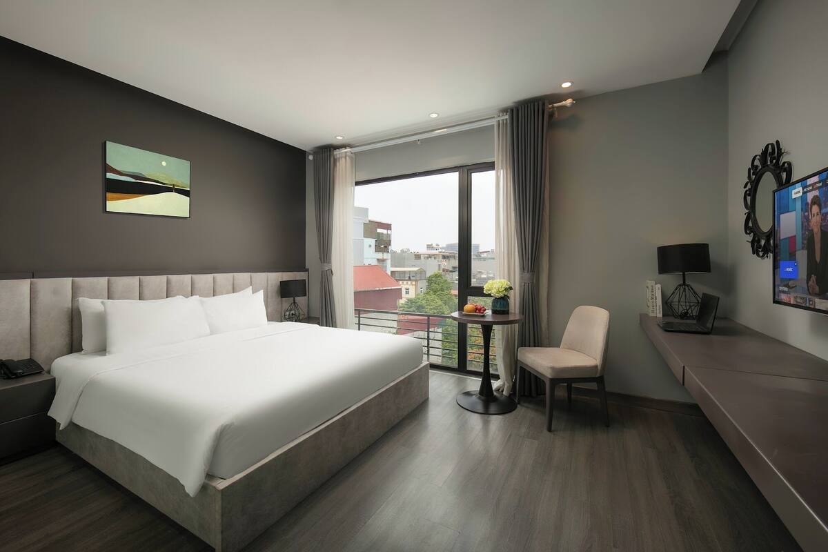 Una habitación de hotel moderna con una cama grande, un escritorio con una computadora portátil, una silla y vistas a la ciudad a través de amplios ventanales.