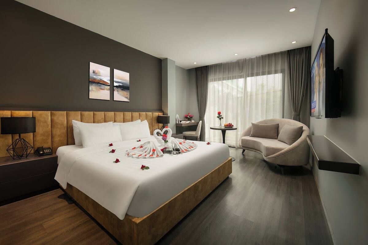 Una habitación de hotel moderna con una cama grande, una mesita de noche con lámpara, una silla, una pequeña mesa con flores y un televisor montado en la pared.
