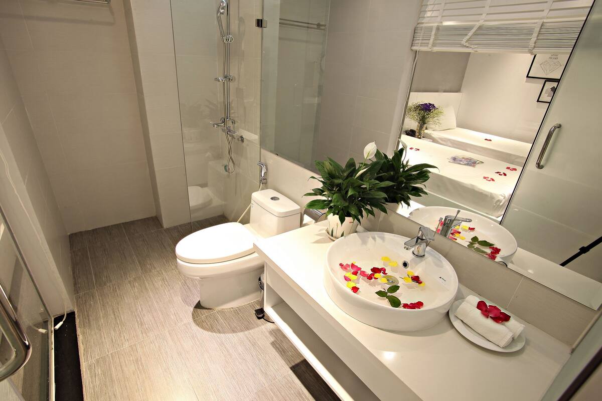 Un baño moderno con cabina de ducha de cristal, inodoro, lavamanos con pétalos de rosa y un espejo que refleja la habitación.