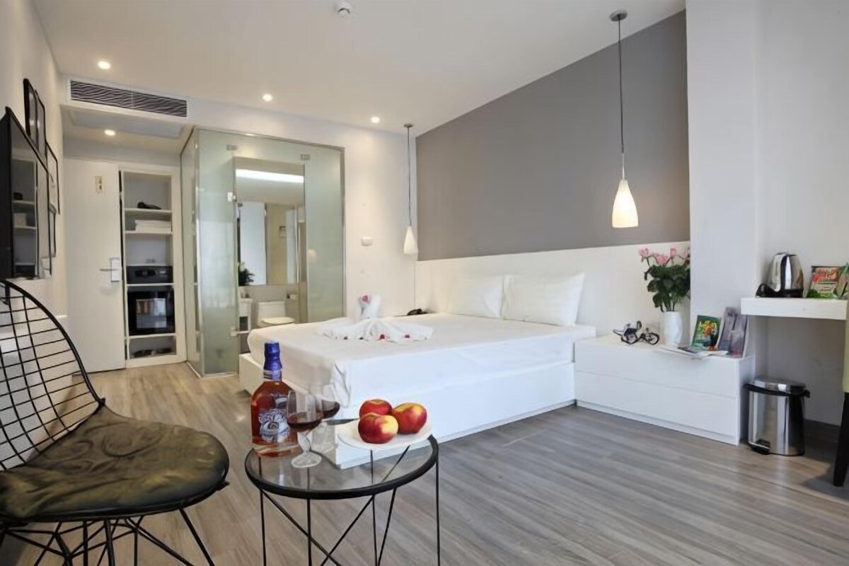 Une chambre d’hôtel moderne avec un lit, une petite table sur laquelle se trouvent des fruits et une bouteille, une chaise, un bureau et un miroir.