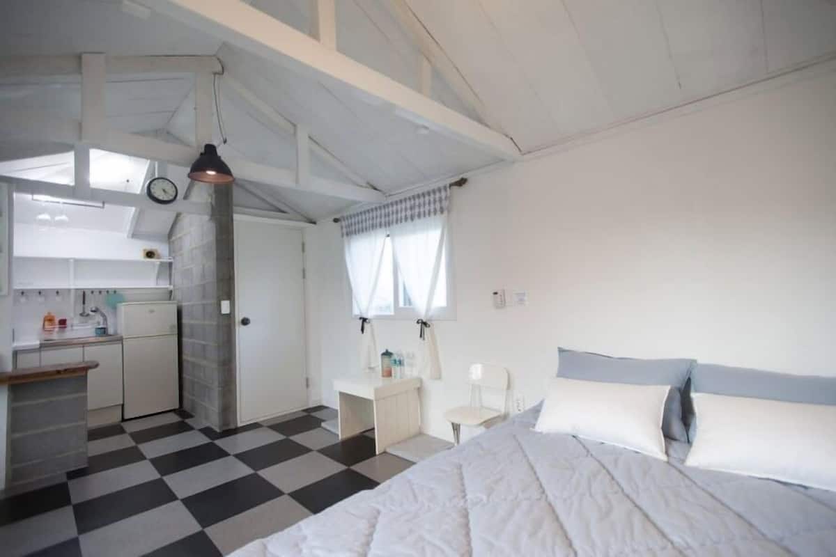 A compact hotel room with a checkered floor, a bed with white and blue bedding, a small kitchenette, and a window with curtains.