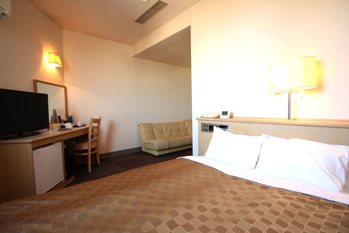 A hotel room with a bed, a desk with a television, a chair, a sofa, and a lamp.