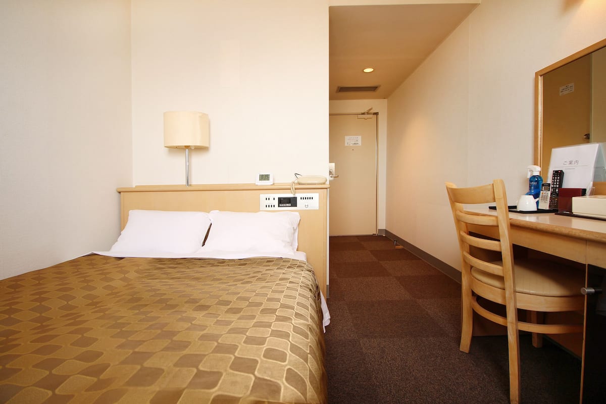 A hotel room with a bed, a desk, a chair, a lamp, a phone, and a mirror.