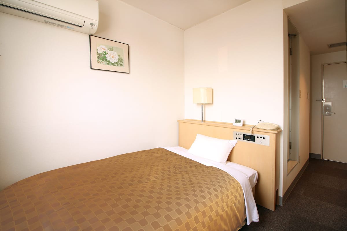 A hotel room with a bed, a bedside table with a lamp, a framed picture on the wall, and an air conditioner.
