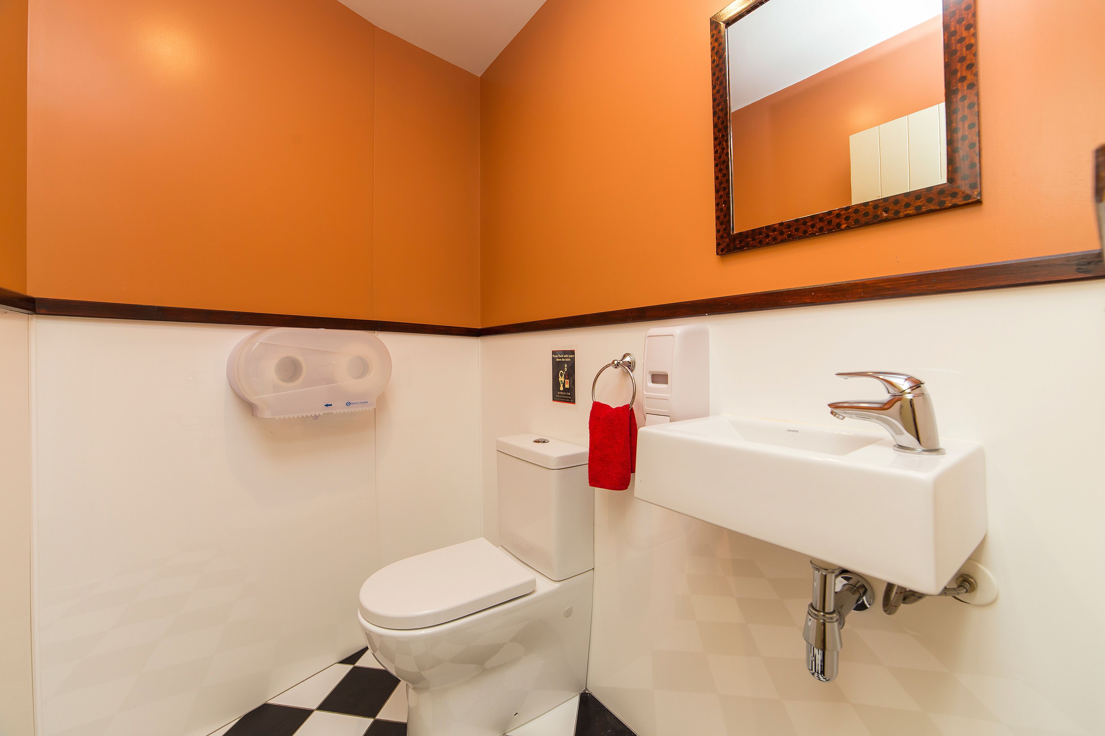 Ein Badezimmer mit orangefarbener Wand, einer weißen Toilette, einem weißen Waschbecken mit Chromarmatur und einem Spiegel mit dekorativem Rahmen.