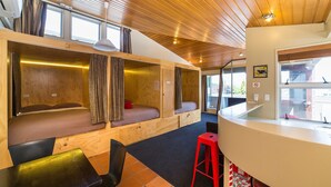 A compact, modern room with built-in seating areas, a small dining table, and a red stool. - Tahuna Pod Hostel (Queenstown)