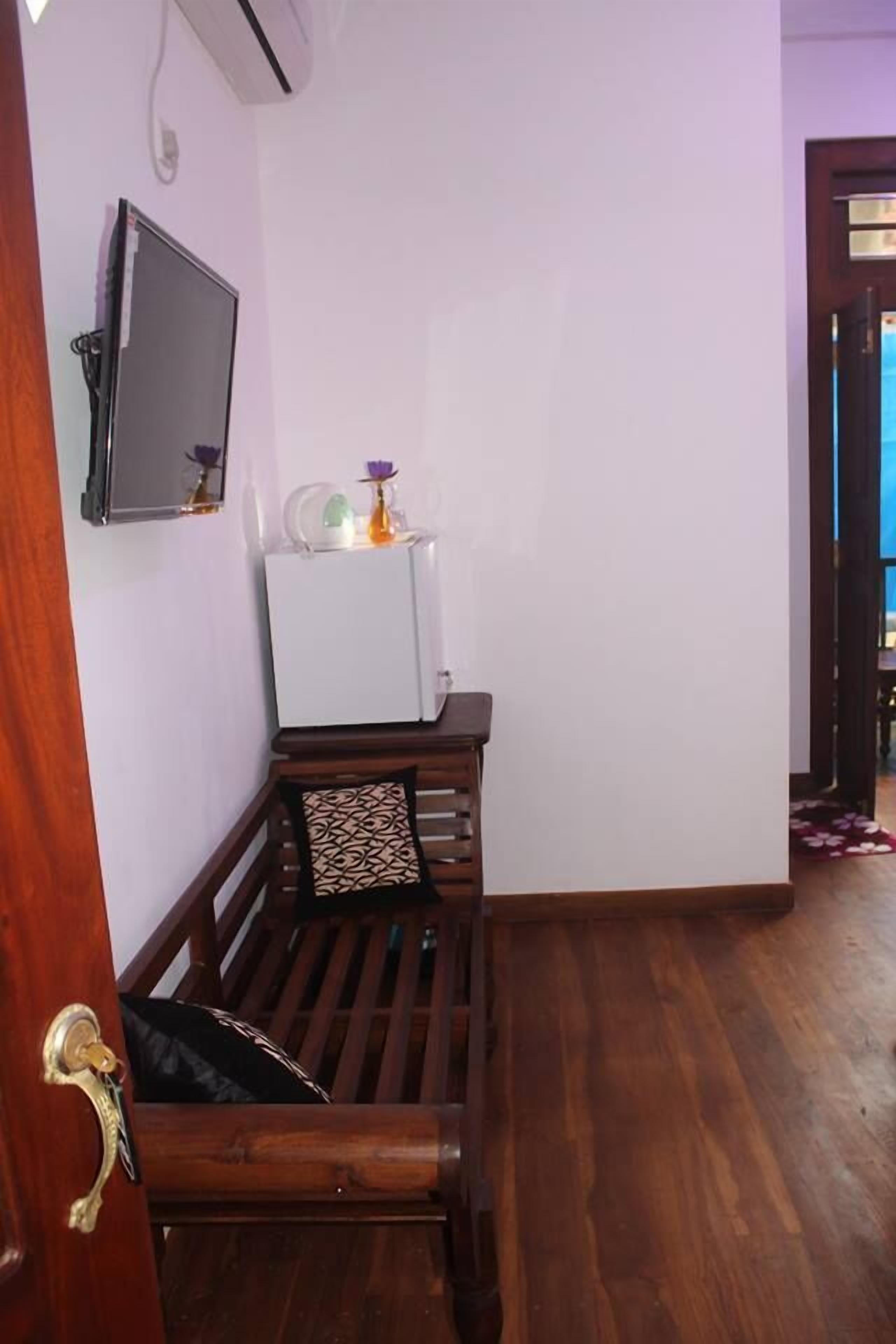 A compact room with a wooden bench, a white refrigerator, a flat-screen TV mounted on the wall, and a door leading to another area.