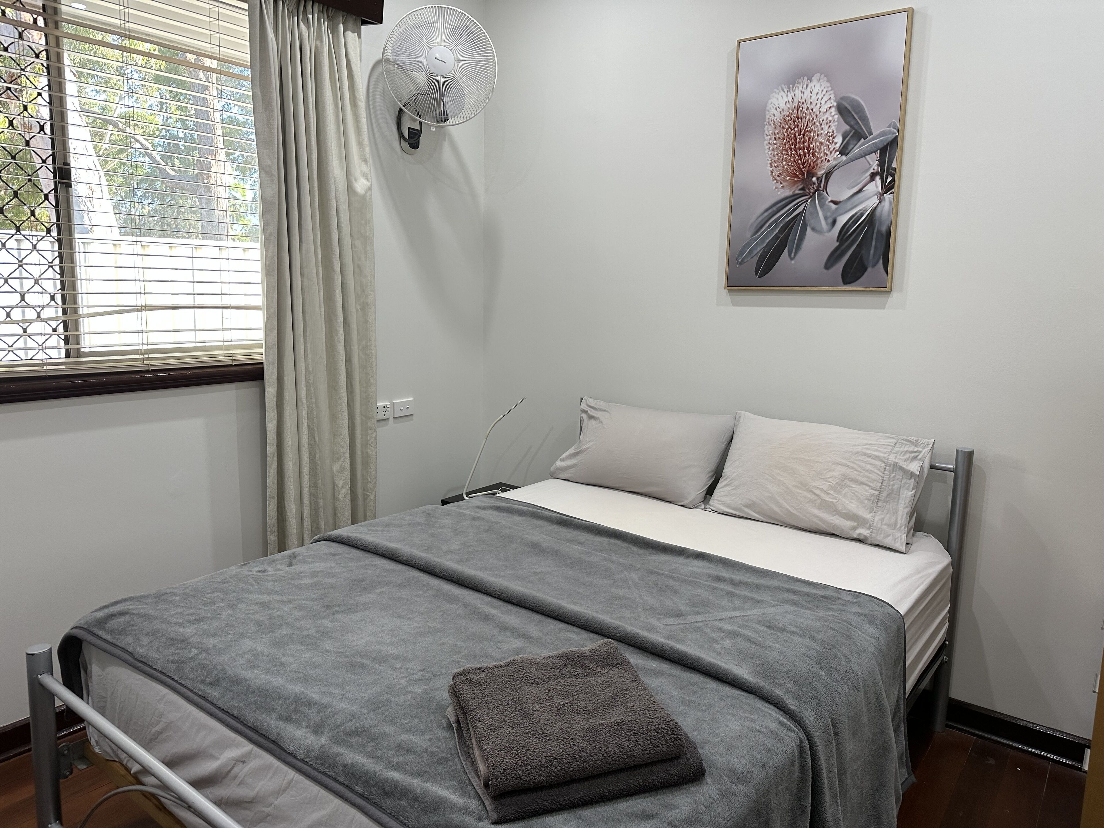 A neatly made bed with a grey blanket and pillows, a standing fan, a window with curtains, and a framed picture on the wall.
