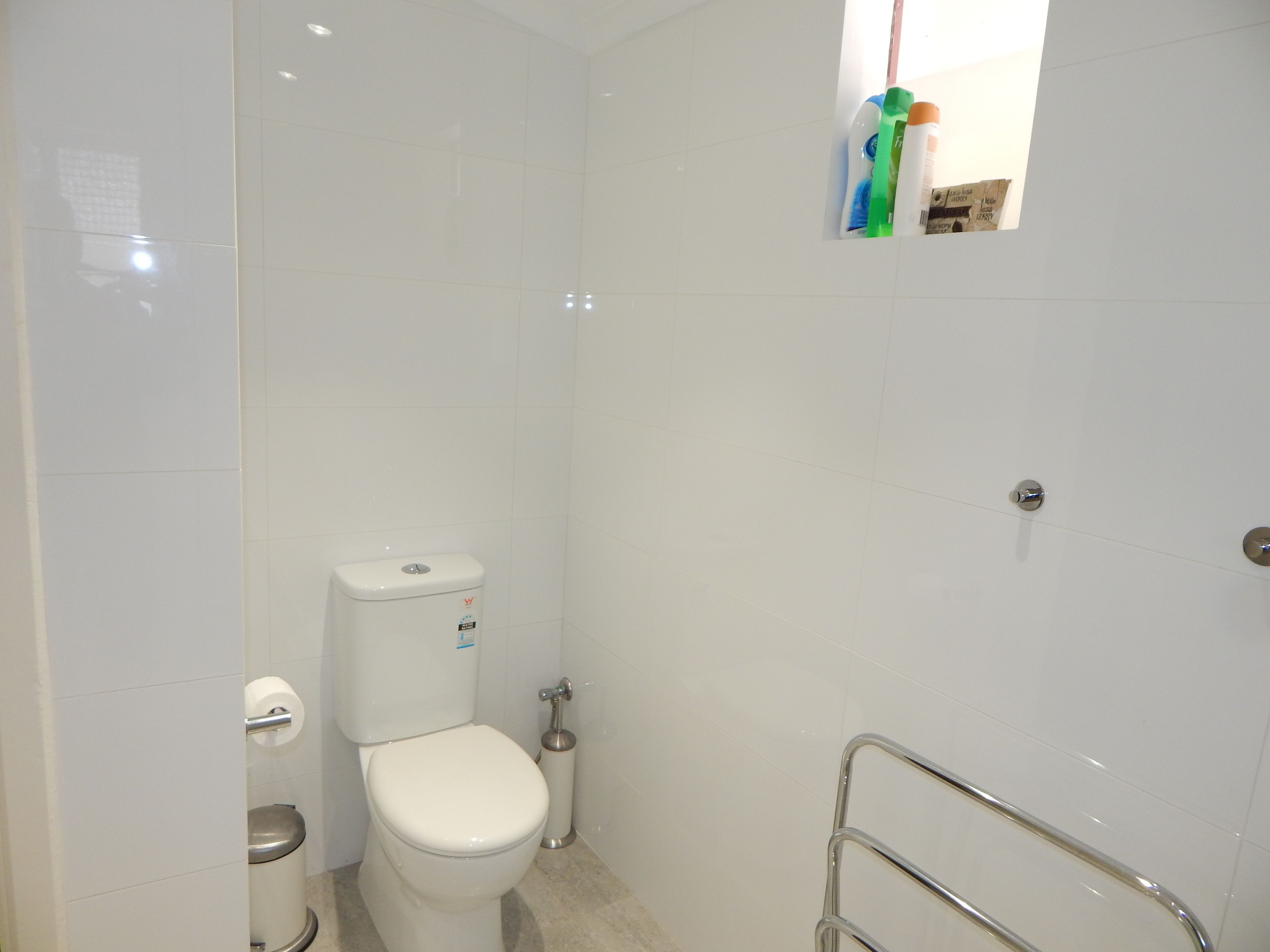A small, clean bathroom with white tiles, a toilet, a hand dryer, and a small shelf with toiletries.