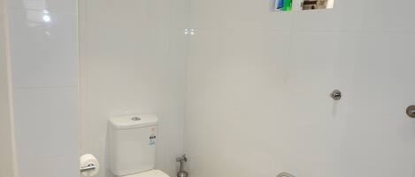 A small, clean bathroom with white tiles, a toilet, a hand dryer, and a small shelf with toiletries.
