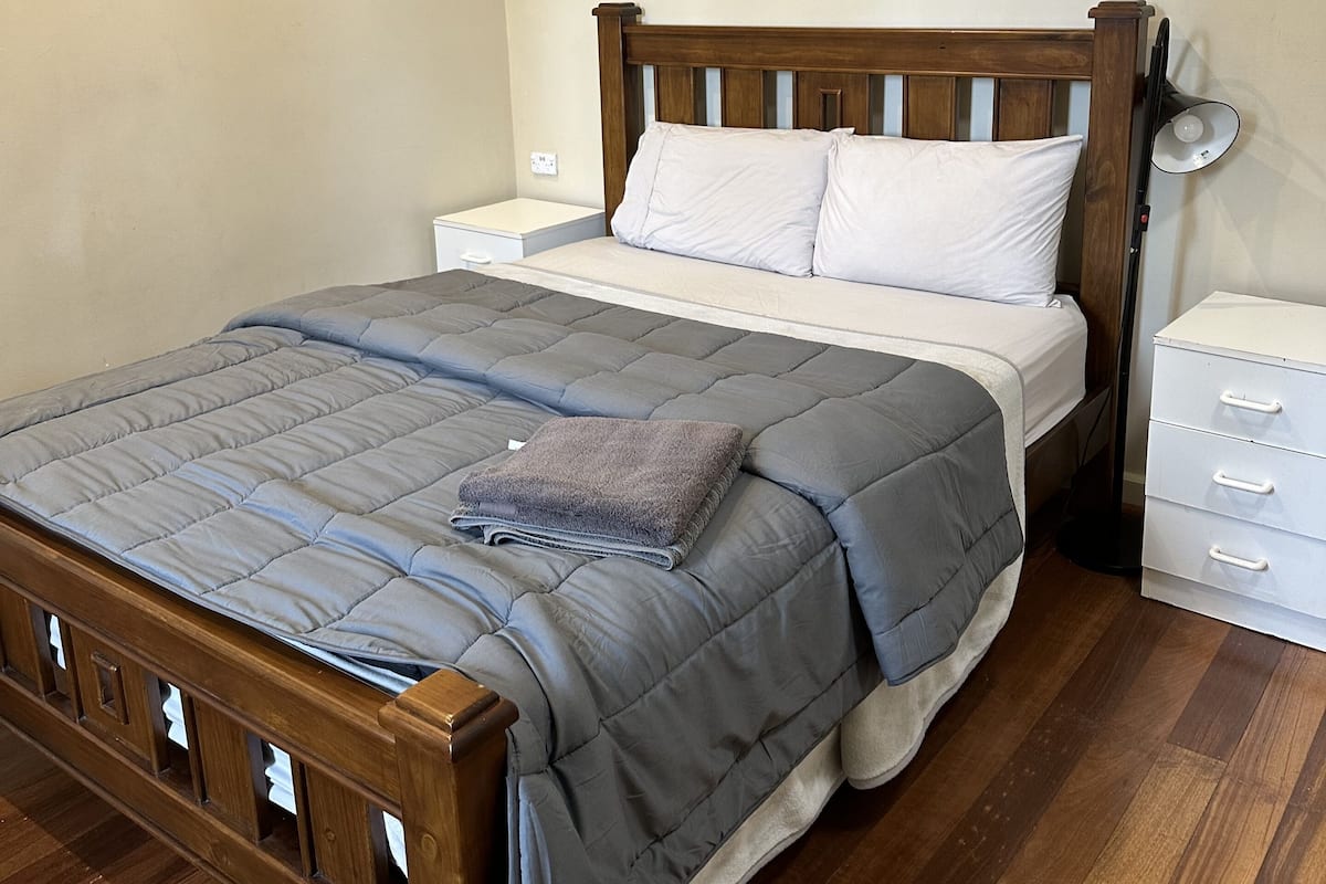 Une chambre à coucher avec un lit en bois, une couette grise, un oreiller blanc, une lampe de chevet et une table de chevet blanche.