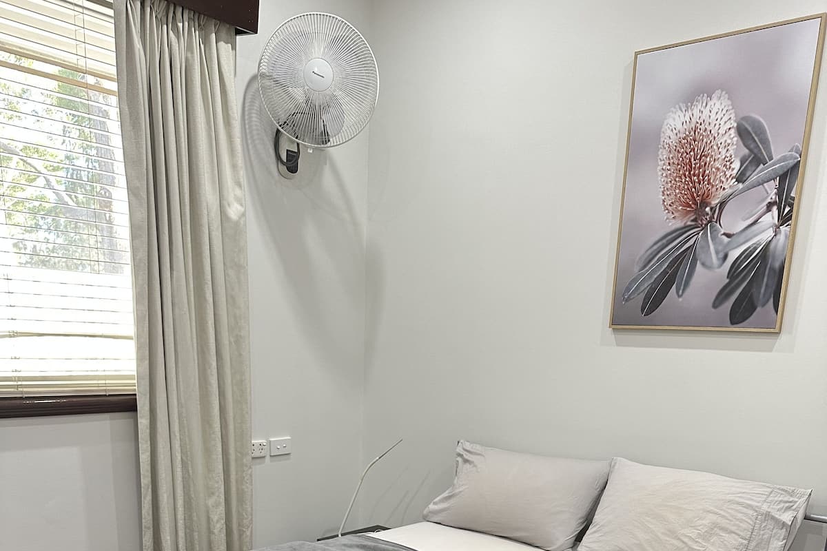 Un lit bien fait, avec une literie grise, une table de chevet avec une lampe, un ventilateur fixé au mur et un tableau représentant une fleur.