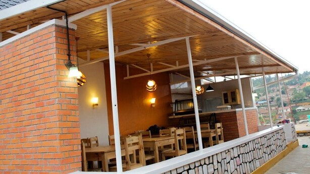 A covered outdoor seating area with brick walls, wooden tables, and chairs, and a view of the surrounding landscape. - Mama Rwanda Youth Hostel (Kigali)