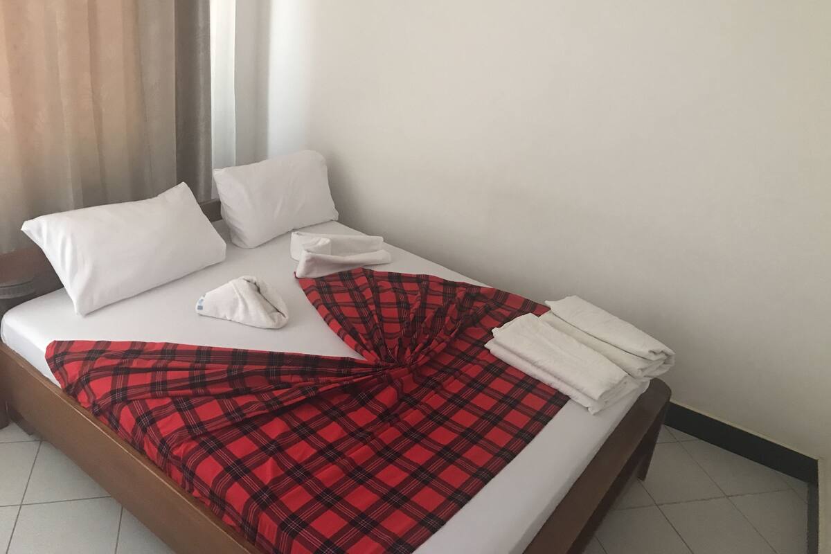 A neatly made bed with white pillows and a red plaid blanket, a window with curtains, and a plain white wall.