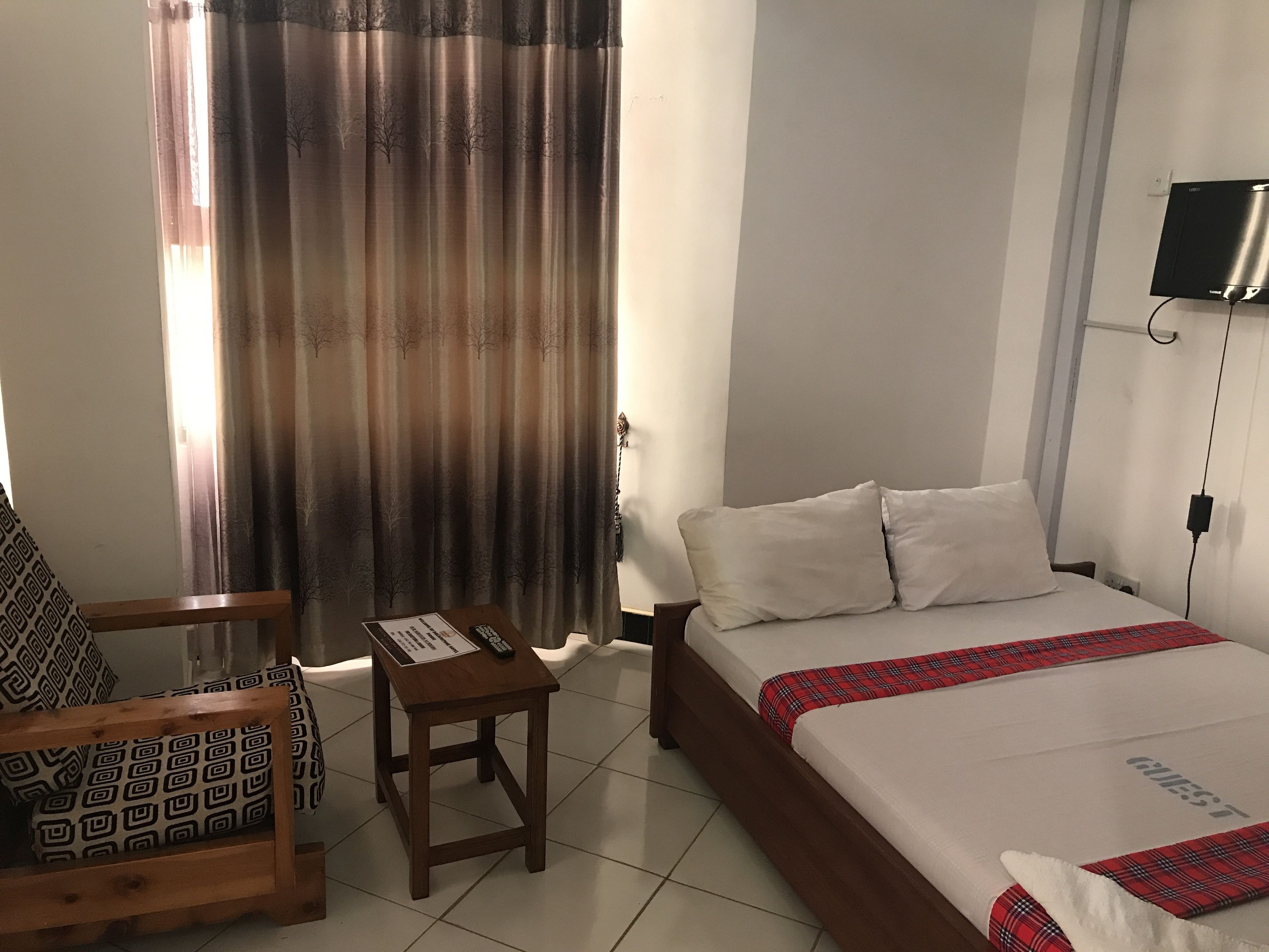 A hotel room with a bed, a wooden chair, a small table with a remote control, and a TV mounted on the wall.