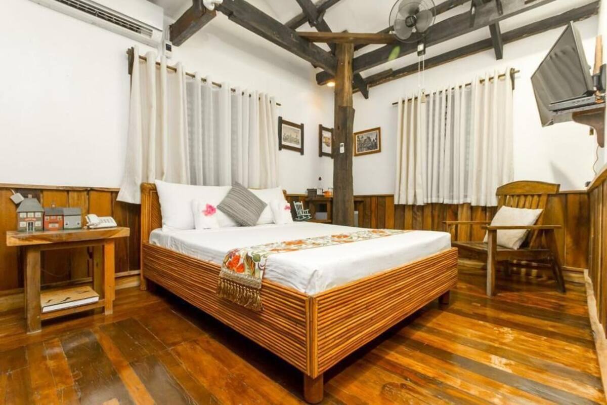 A wooden-framed bed with white bedding, a small bedside table with a lamp, and a chair with a cushion.