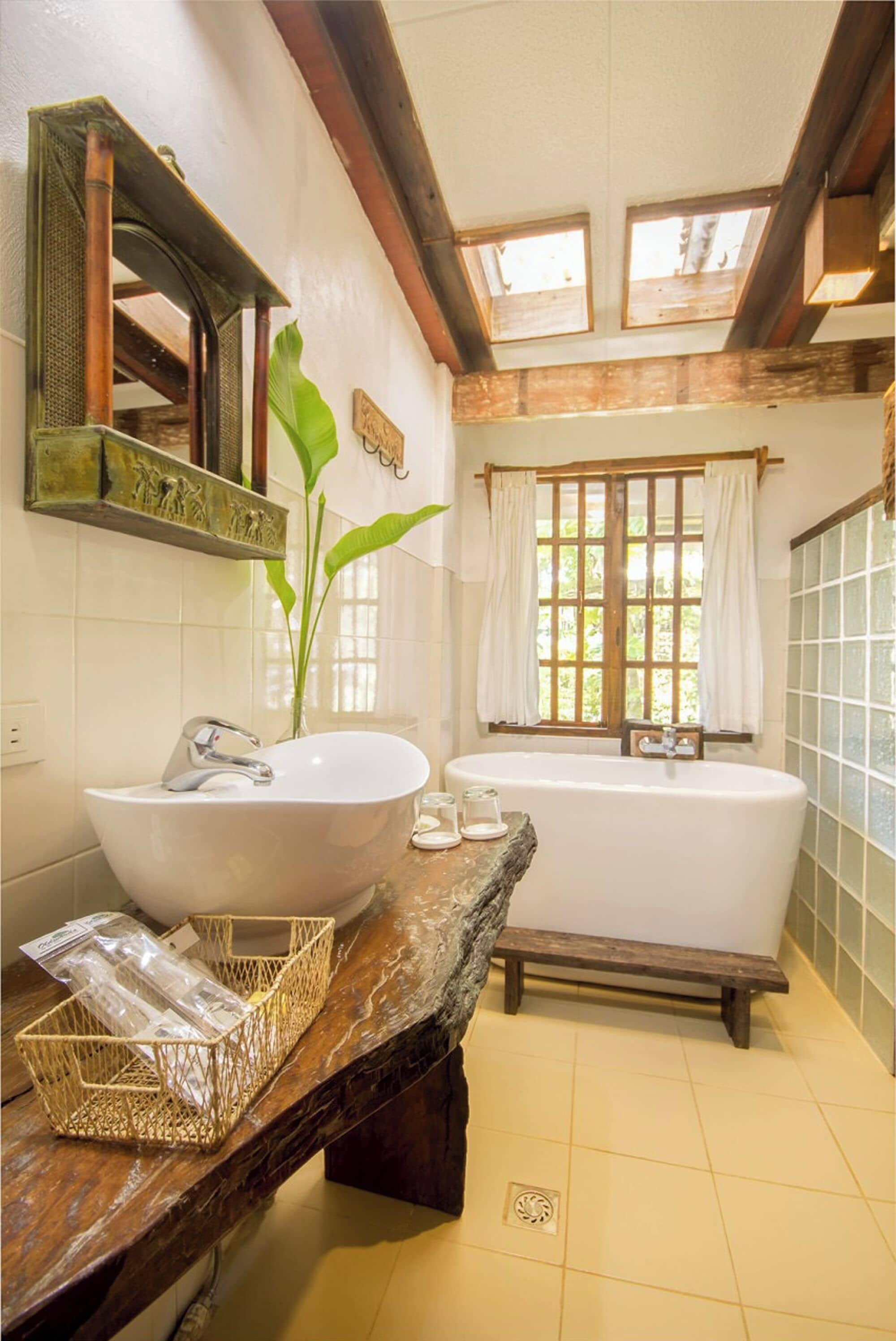 A bathroom with a large white sink, a bathtub, wooden beams, a window with curtains, and a mirror with a decorative frame.
