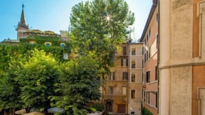 A narrow street with buildings on either side, a tree-lined courtyard, and a distant church spire. - Rome Visits (Rome)