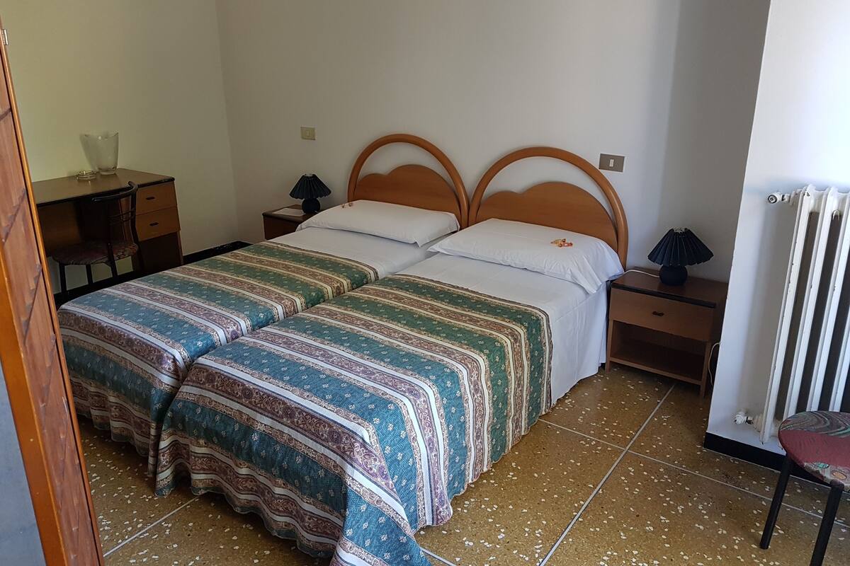 A double bed with a patterned bedspread, a wooden headboard, two bedside tables with lamps, a chair, and a radiator.