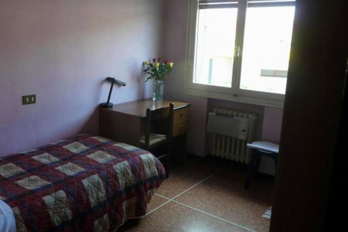 A bedroom with a bed, a desk, a chair, a radiator, and a window.
