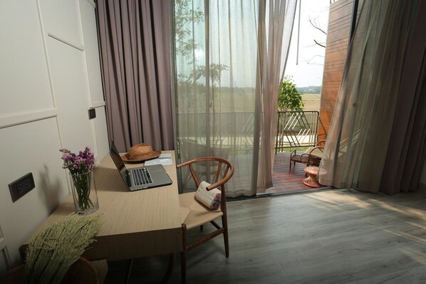 Una habitación con un escritorio de madera, una computadora portátil y un florero. Hay una silla con cojín y vistas al exterior a través de una puerta de cristal.
