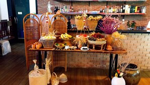 Una mesa de madera con diversas frutas, cestas y objetos decorativos en una habitación con paredes de ladrillo y una zona de bar al fondo.