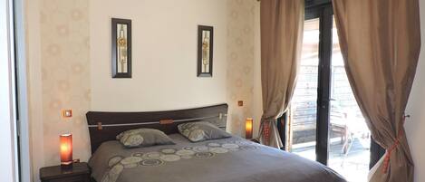 A bedroom with a large bed, two wall-mounted picture frames, a bedside table with a lamp, and a view of the outside through a window with curtains.