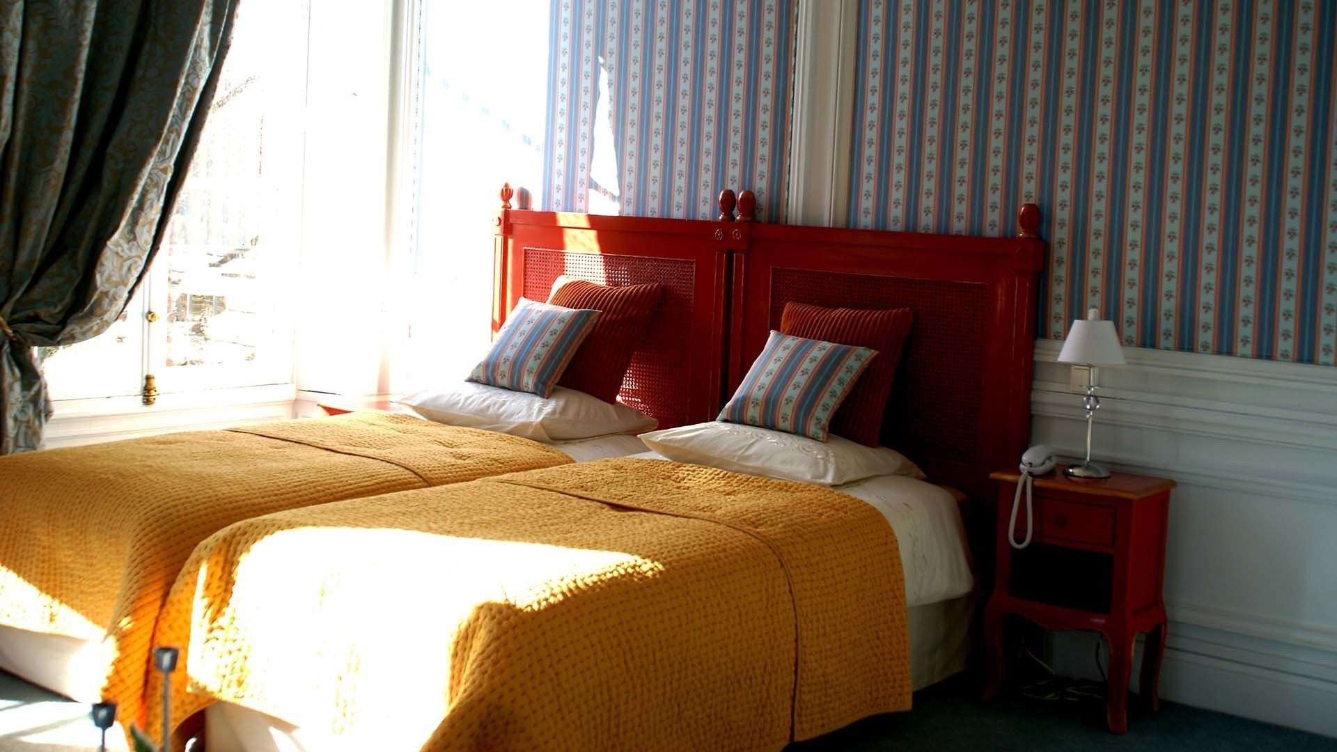 A double bed with a red headboard, two pillows, and a yellow blanket. There is a bedside table with a lamp and a phone. The room has striped wallpaper and a window with curtains.