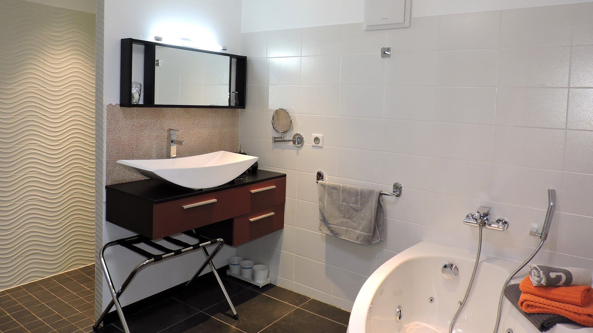 Un baño moderno con un lavamanos blanco, tocador de madera oscura, un espejo grande, un perchero para toallas y una bañera con ducha.