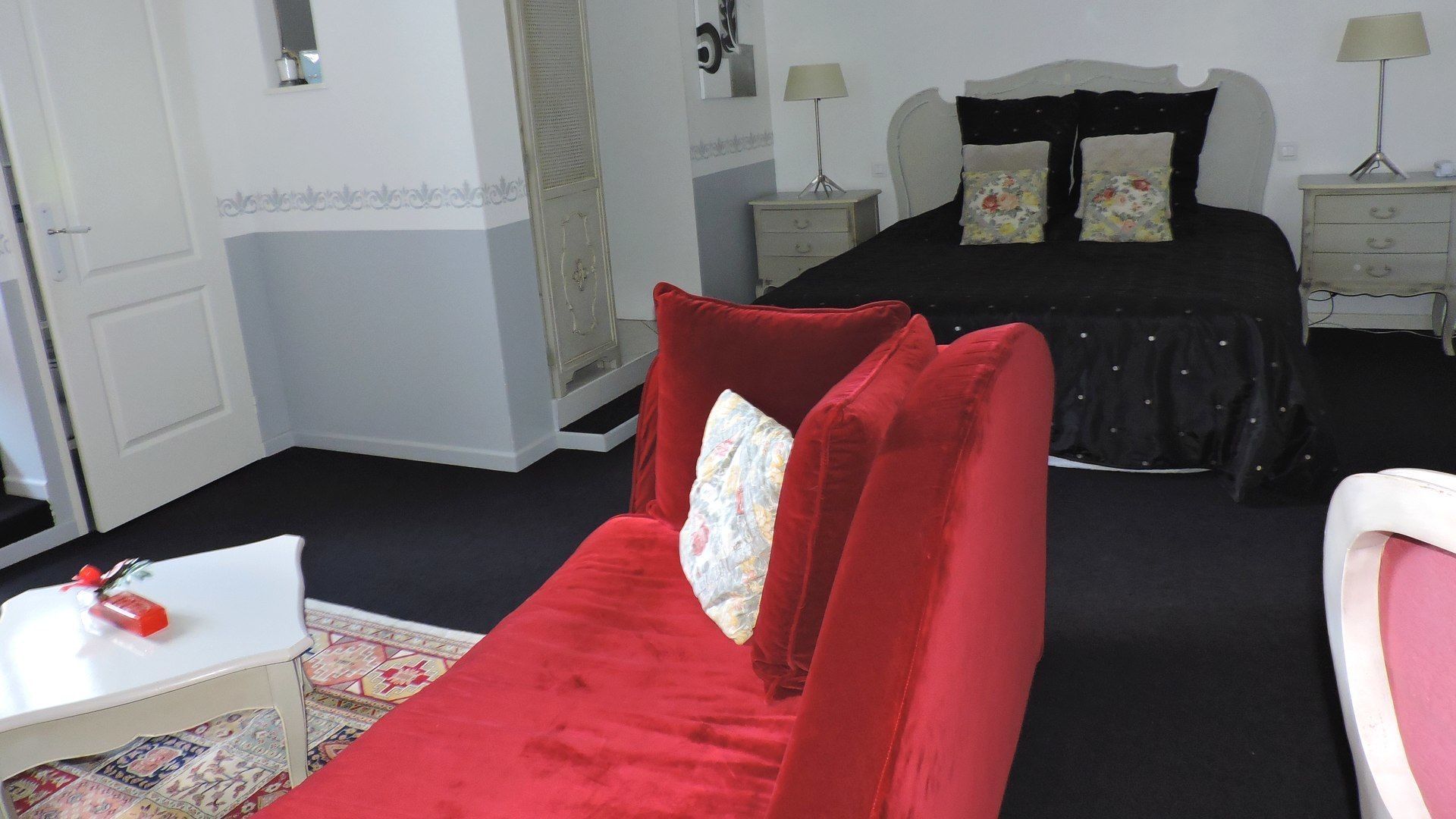 A bedroom with a red sofa, a black bed with floral pillows, a white bedside table, and a nightstand with a lamp.