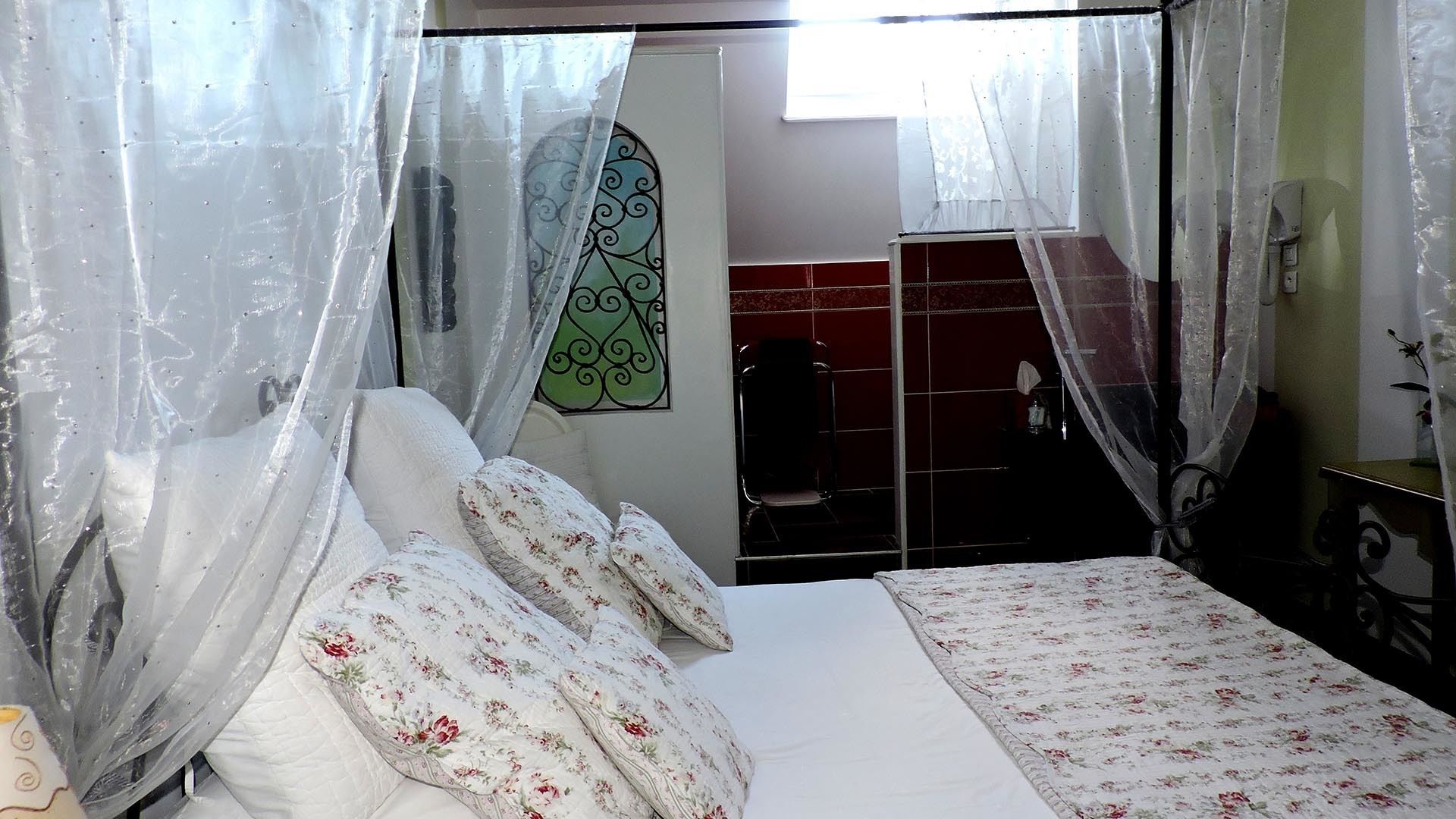 A bedroom with a bed, floral bedding, a window with sheer curtains, and a red tiled wall.