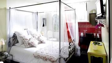 A four-poster bed with a canopy, a bedside table with a lamp, a small table with a plant, and a bathroom with a red-tiled wall.