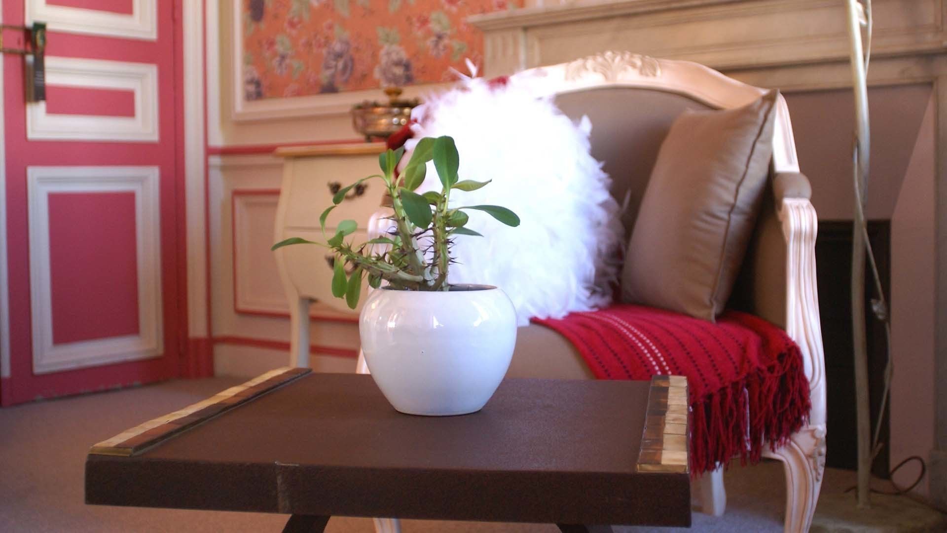 A room with a white vase on a small table, a beige armchair with a red throw blanket, and a pink wall with floral wallpaper.
