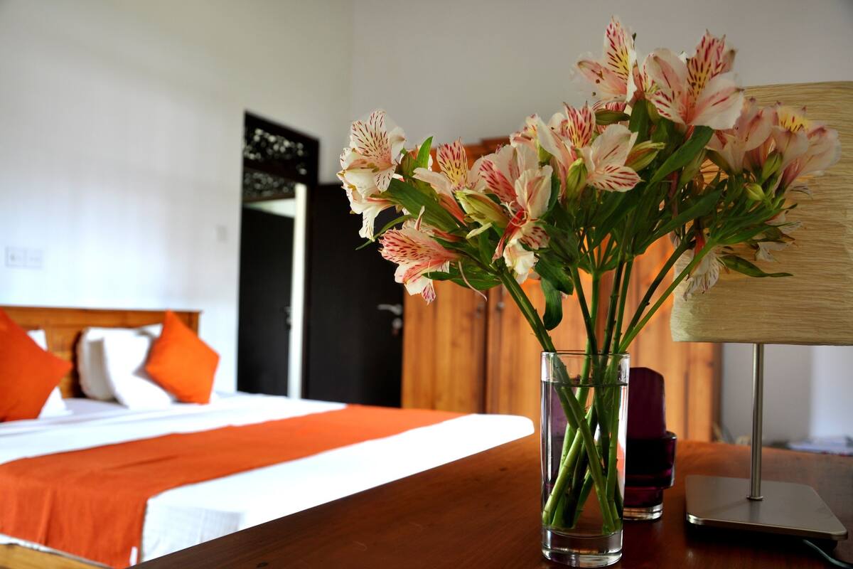 Habitación de hotel con cama, ropa de cama naranja, un jarrón de flores en la mesita de noche y una lámpara.