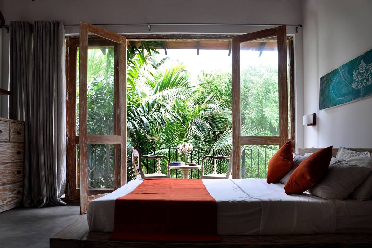 Un dormitorio con un ventanal amplio, una cama con almohadas, un cabecero de madera y vistas a la vegetación exuberante.