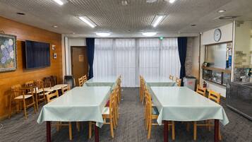 A conference room with two long tables, wooden chairs, a clock, and a kitchen area with a sink and stove.