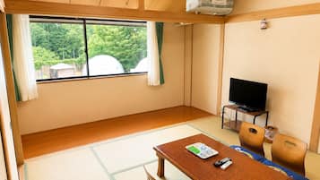 A room with a television, a table with remote controls, and a view of greenery outside.