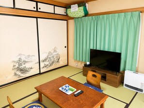 Une pièce de style japonais traditionnel, avec un sol recouvert de tatamis, un téléviseur, une table en bois avec une télécommande et un rideau vert.