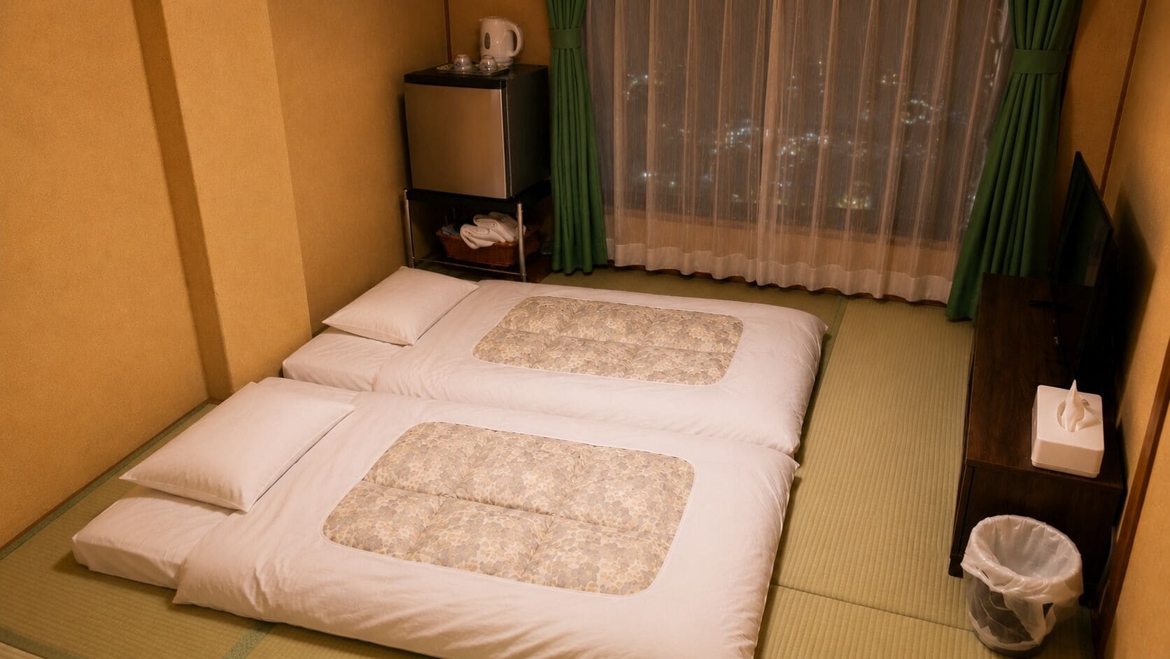 Hotelkamer met twee bedden, een kleine tafel met waterkoker, een prullenbak en uitzicht op de stad 's nachts.