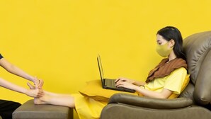 A person sitting on a leather couch with a laptop, wearing a yellow dress and a brown scarf, against a yellow background. - 128 Room And Massage (Bangkok)