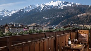 Un balcón de madera con una mesa, una botella y vasos, con vistas a una cadena montañosa y un pueblo.
