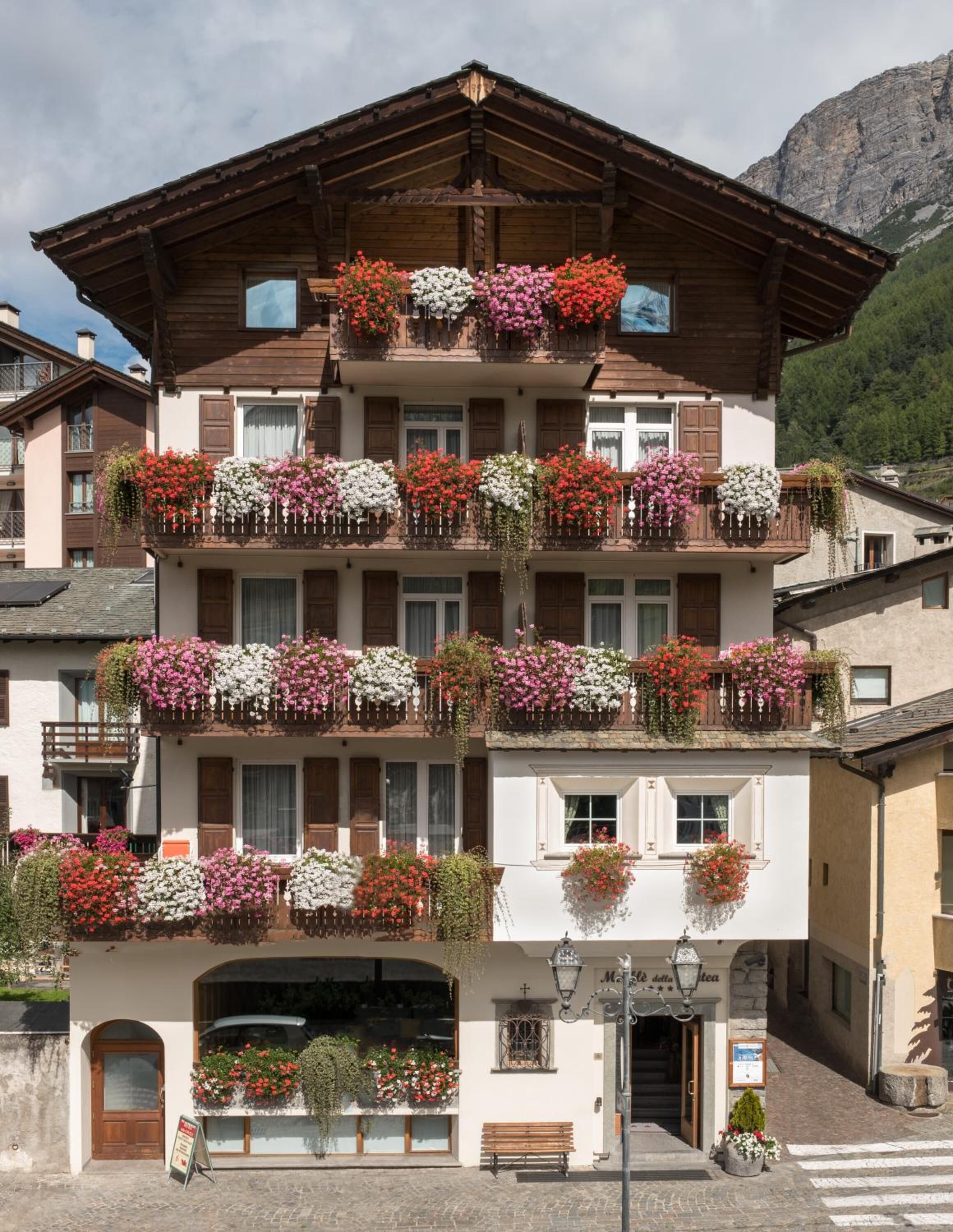 A multi-story building with balconies adorned with flowers, a ground-level entrance, and a mountain backdrop.