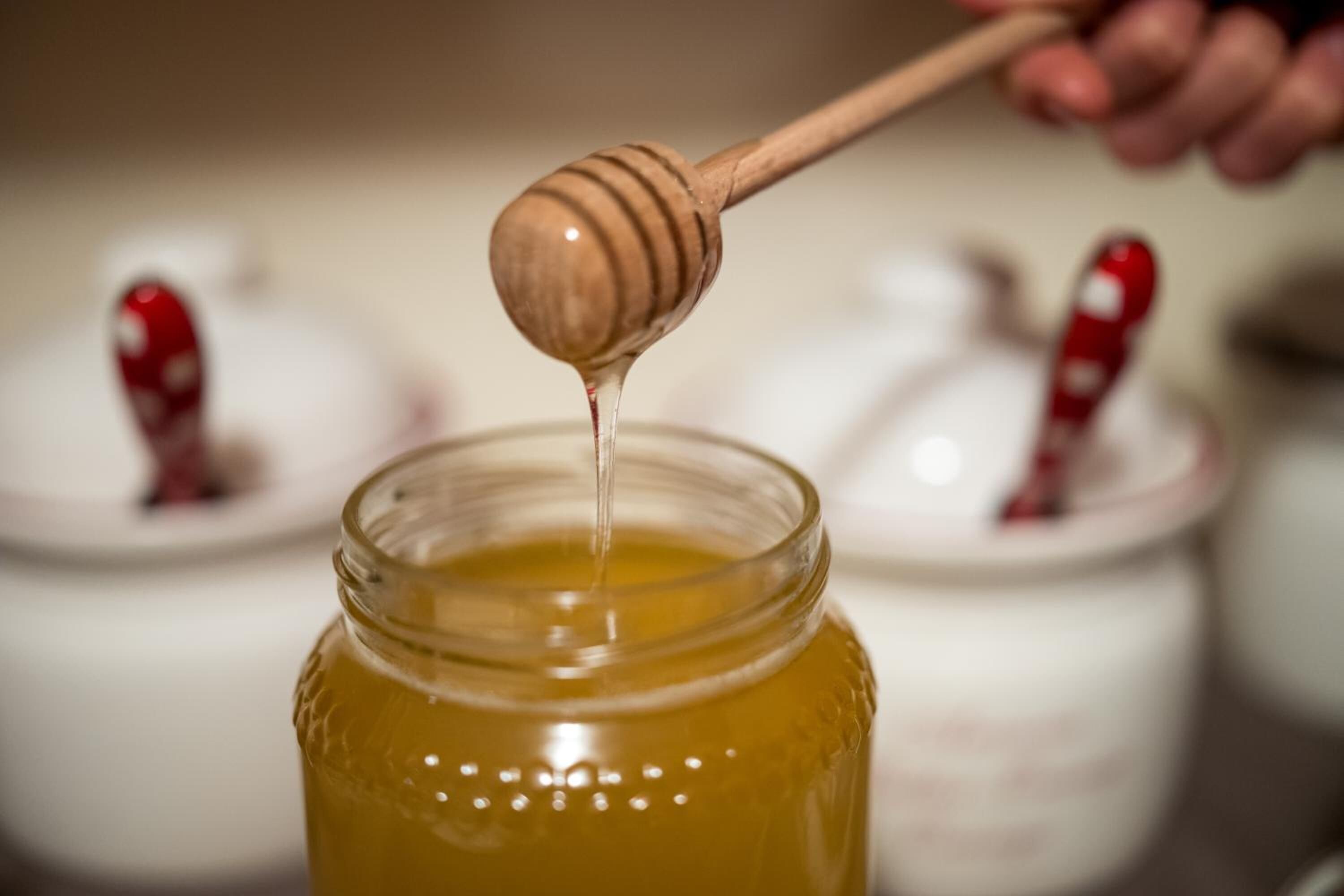 A close-up of a honey jar with a wooden honey dipper dripping honey into it.
