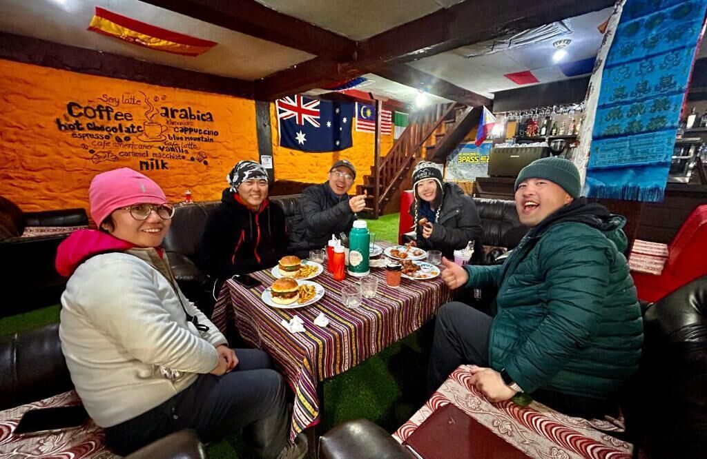 A group of people seated around a table with food and drinks, in a room with a wall displaying various coffee and tea options.