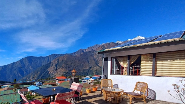Une terrasse meublée de mobilier en osier, avec une table et des chaises, donnant sur une chaîne de montagnes où l’on aperçoit des panneaux solaires sur un bâtiment.
