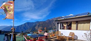 A terrace with wicker furniture, a table, and chairs, overlooking a mountain range with solar panels on a building. - Lama Hotel (Lukla)