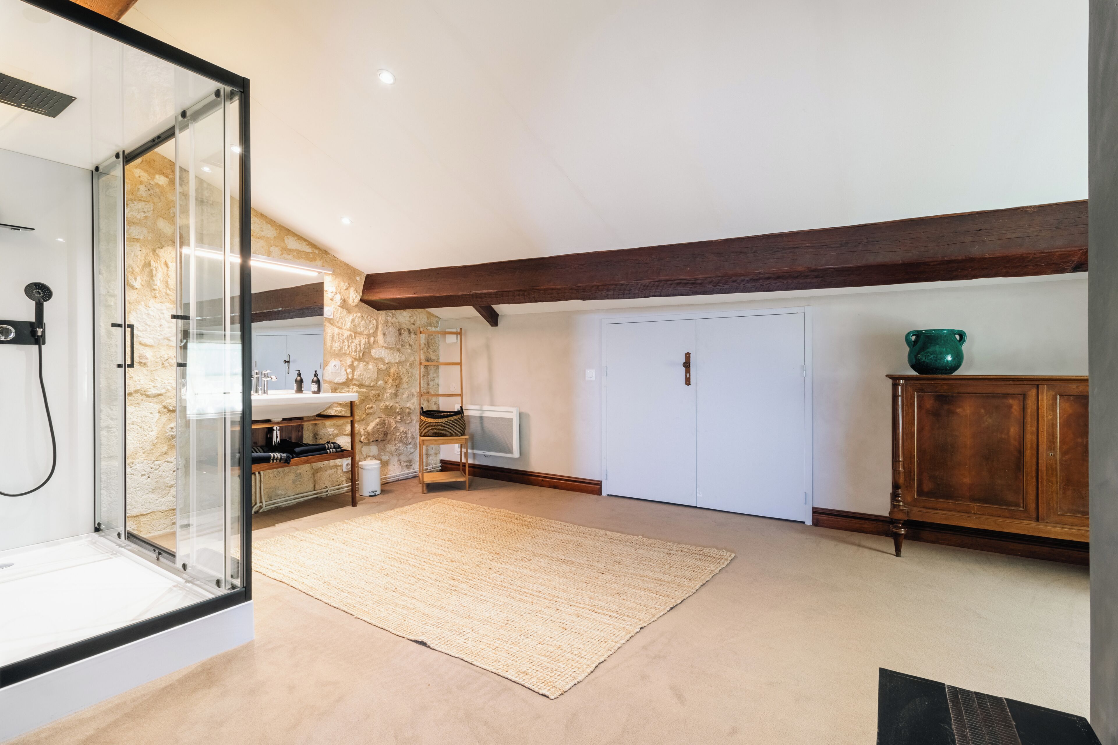 Un baño moderno con cabina de ducha de vidrio, pared de piedra, viga de madera en el techo, lavabo blanco y mueble de madera.